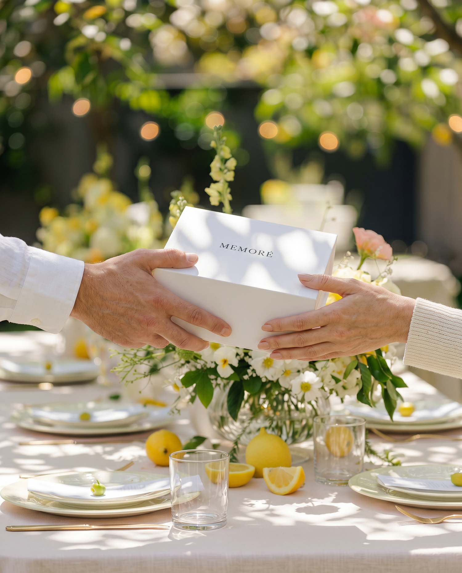 Two hands exchanging a 'MEMORE' giftbox at an outdoor birthday party floral and lemon decorations.